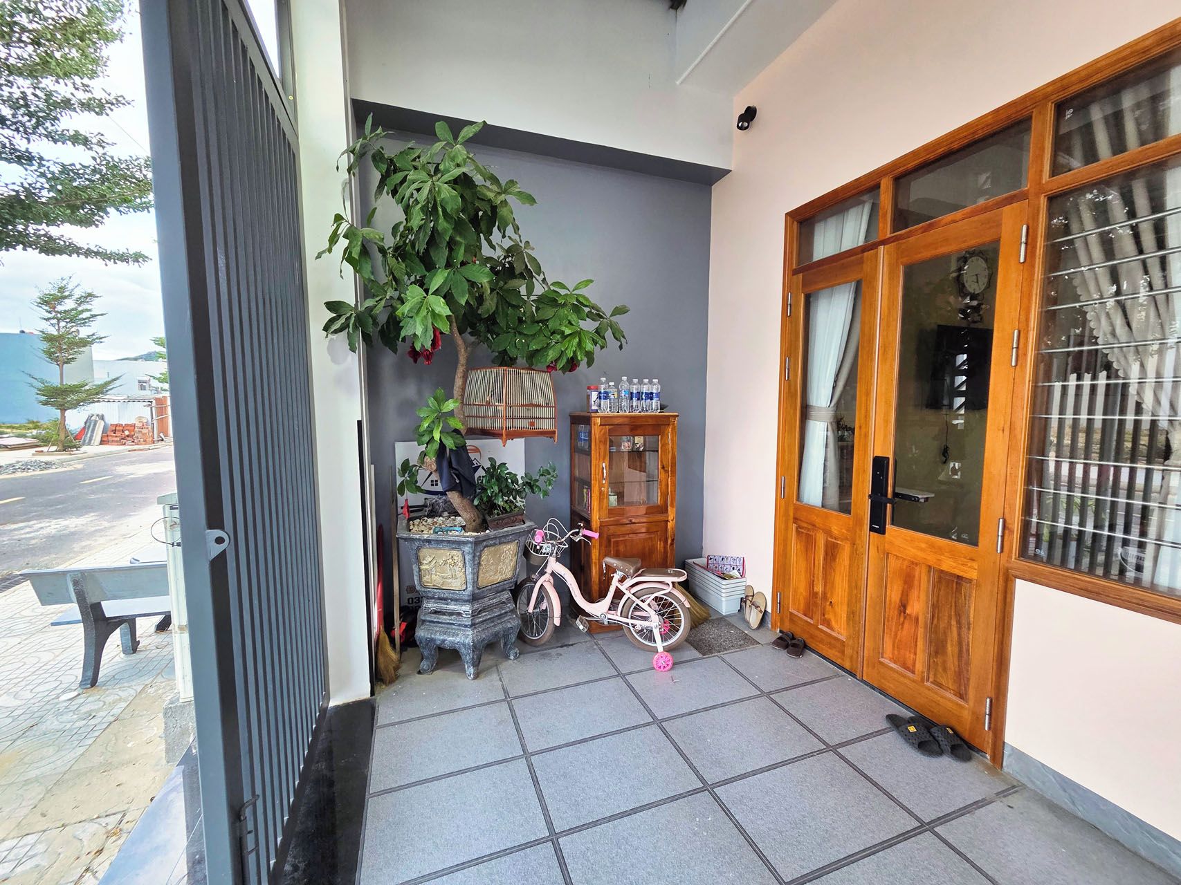 Entrada d'un edifici residencial amb un porxo enrajolat, una bicicleta, un armari de fusta i una planta en test.