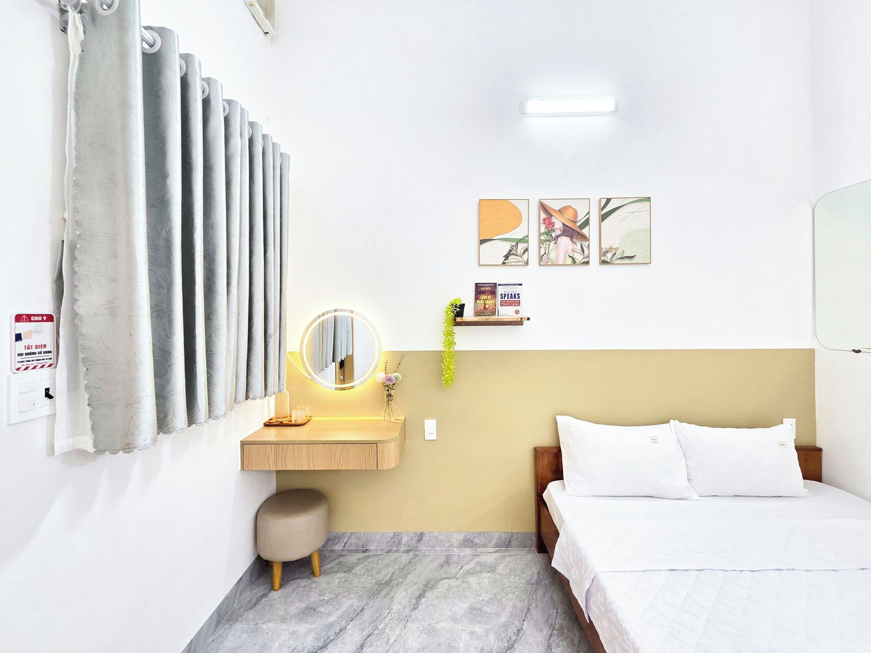 A hotel room with a bed, a bedside table, a mirror, a wall-mounted shelf with books, and a window with curtains.