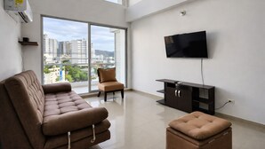Un salon moderne avec un canapé, un pouf et un téléviseur, de grandes fenêtres offrant une vue sur la ville, et un téléviseur à écran plat fixé au mur.