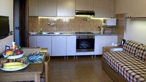 Une cuisine compacte avec des meubles blancs, un four à micro-ondes, une cuisinière et un évier. On y trouve une table à manger avec des fruits et un canapé à carreaux.
