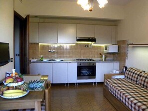 Eine kompakte Küche mit Mikrowelle, Herd und Backofen. Ein Esstisch mit Obst und ein kariertes Sofa sind ebenfalls zu sehen.