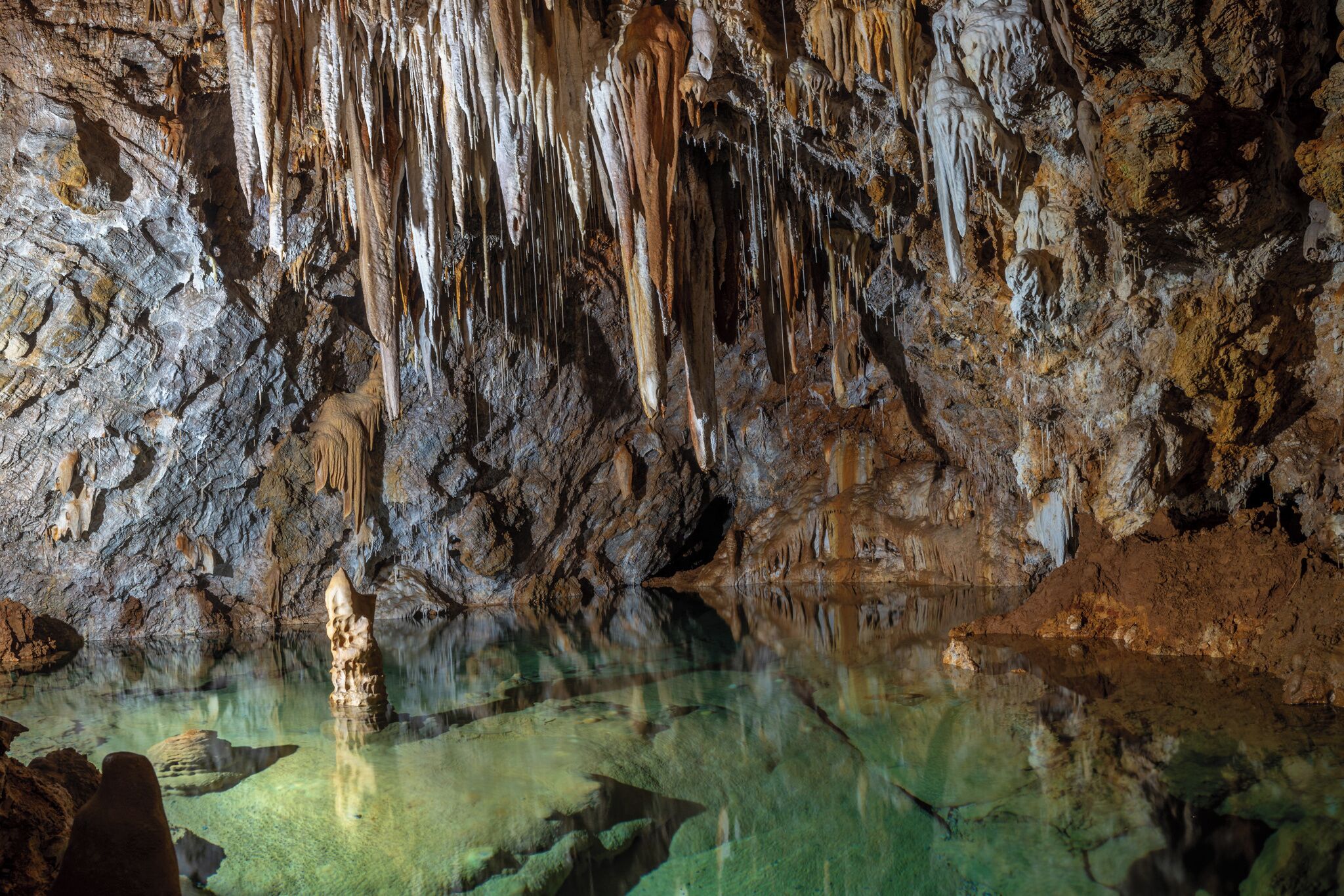 Eine Höhle mit Stalaktiten und Stalakmitten, klarem Wasser und einem steinigen Boden.