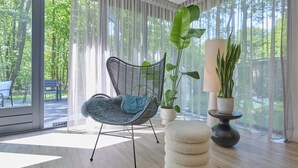 A modern room with a large window, a wicker chair, a round ottoman, and two potted plants.