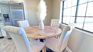 A dining area with a round table, four upholstered chairs, a vase with white feathers, a chandelier, and a large window with blinds. - Nice Modern Vintage House near Sky Harbor Airport (Phoenix)