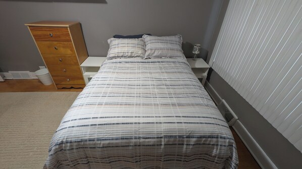 A single bed with a striped bedspread, a wooden dresser, a nightstand, and a window with blinds.