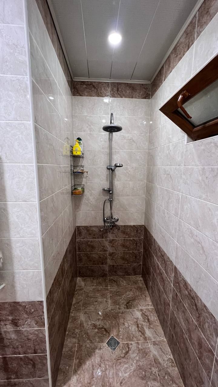 A small, tiled shower room with a showerhead, a handrail, and a shelf with toiletries.
