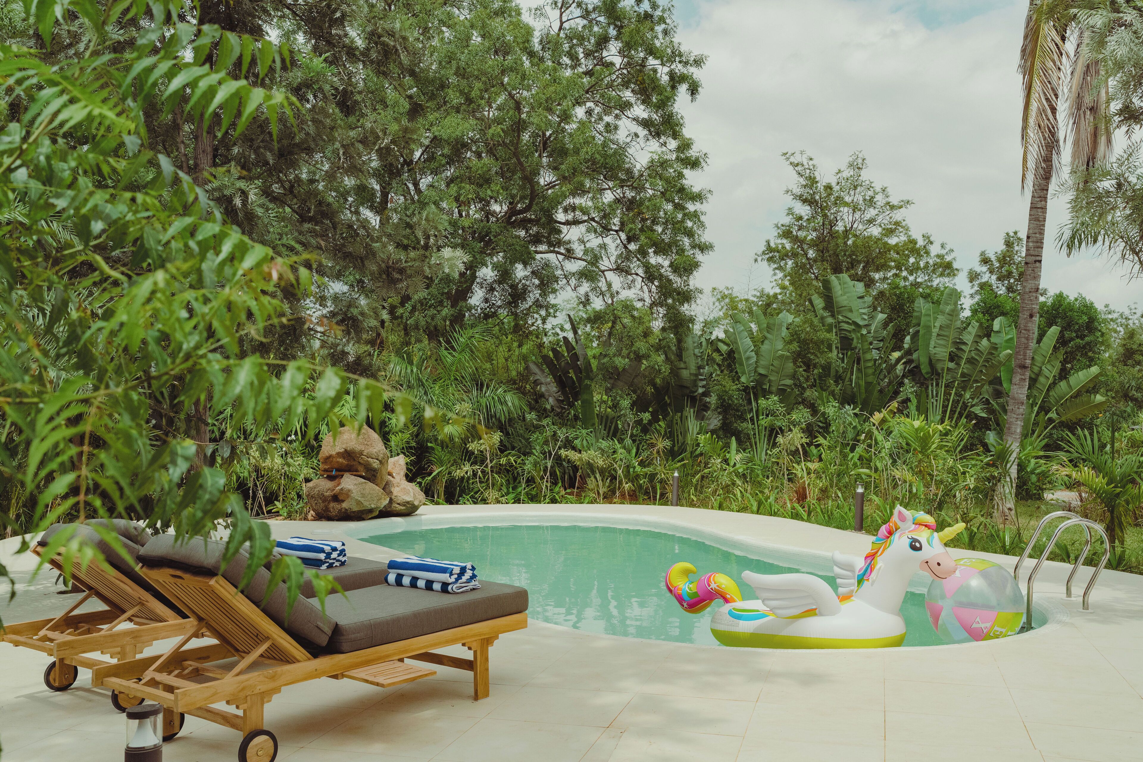 Un área de piscina con sillones de descanso, un flotador de unicornio inflable y abundante vegetación.