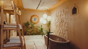 Een badkamer met een stenen muur, een badkuip, een houten ladder, rieten manden en een spiegel met een ronde omlijsting.