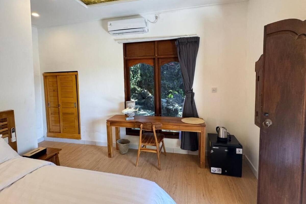 A bedroom with a bed, a wooden desk, a chair, a window with curtains, a wardrobe, and an air conditioning unit.