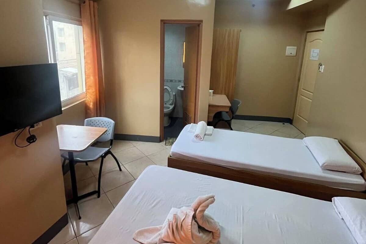 Ein Hotelzimmer mit zwei Betten, einem Schreibtisch, einem Stuhl, einem Fernseher und einem durchgehenden Türbogen einsehbarer Badezimmer.