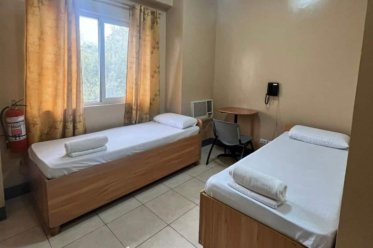 Ein Zimmer mit zwei Einzelbetten, einem kleinen Tisch, einem Stuhl, einem Feuerlöscher und einem Vorhangfenster.