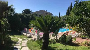 A garden with a stone pathway, a palm tree, and a pool area with colorful lounge chairs. - Holiday apartment mit Klimaanlage und Pool (Loano)