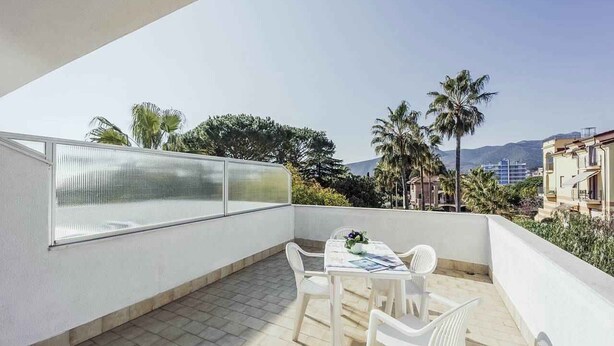 A balcony with a table and chairs, a glass railing, and a view of palm trees and buildings. - Holiday apartment mit Klimaanlage und Pool (Loano)