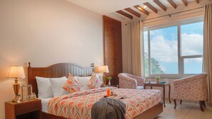 A hotel room with a large bed, two bedside tables, a chair, a small table, and a window with curtains. - Elivaas Konifer Greyvine Hillside 9 BHK  (Kasauli)