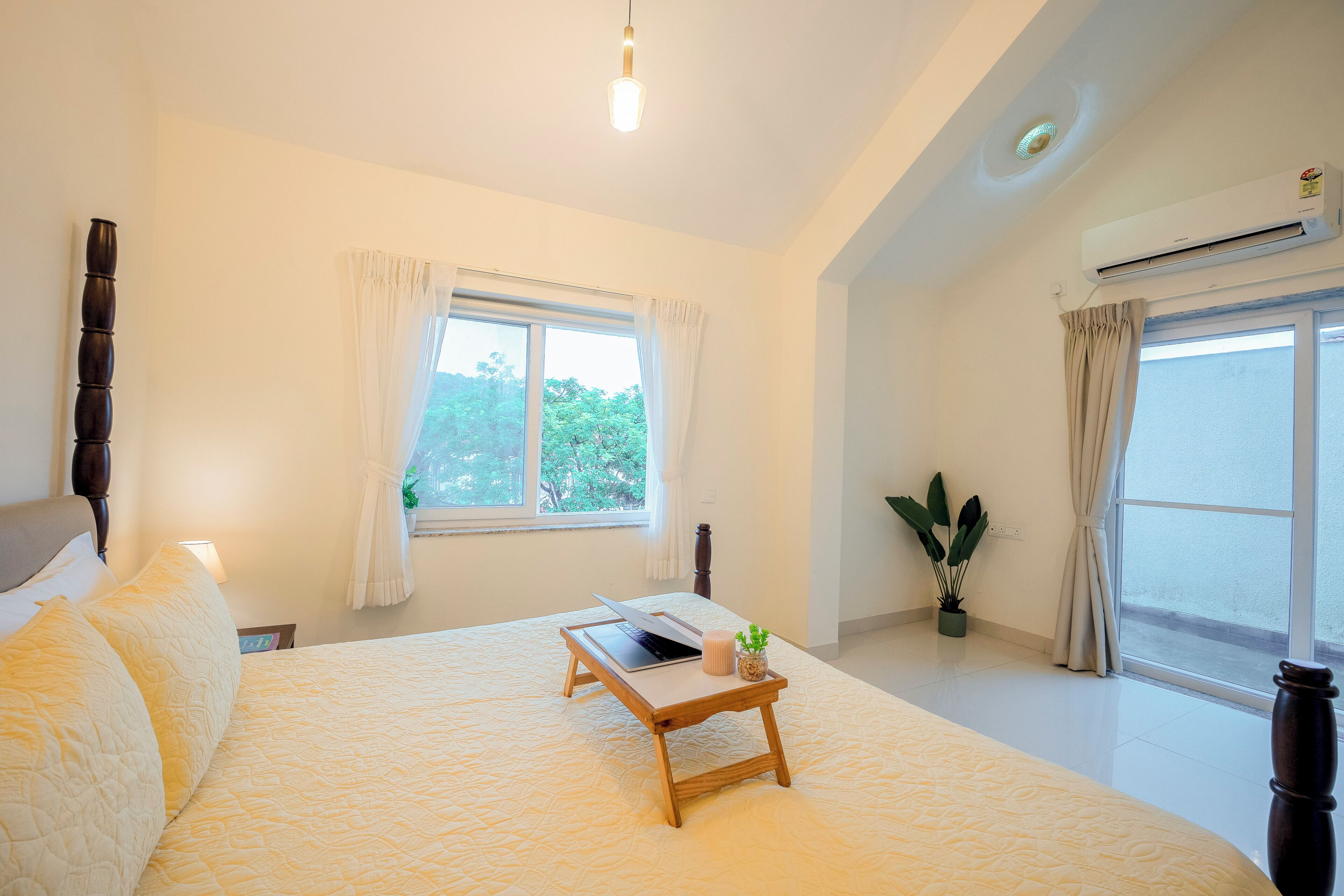 A bedroom with a bed, a bedside table with a laptop, a potted plant, and a window with curtains.