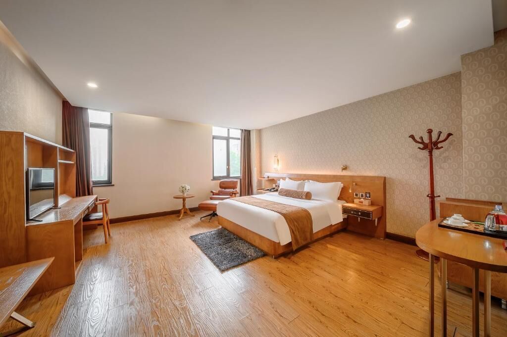 A hotel room with a bed, desk, chair, bedside table, lamp, coat rack, and a view of the outside.