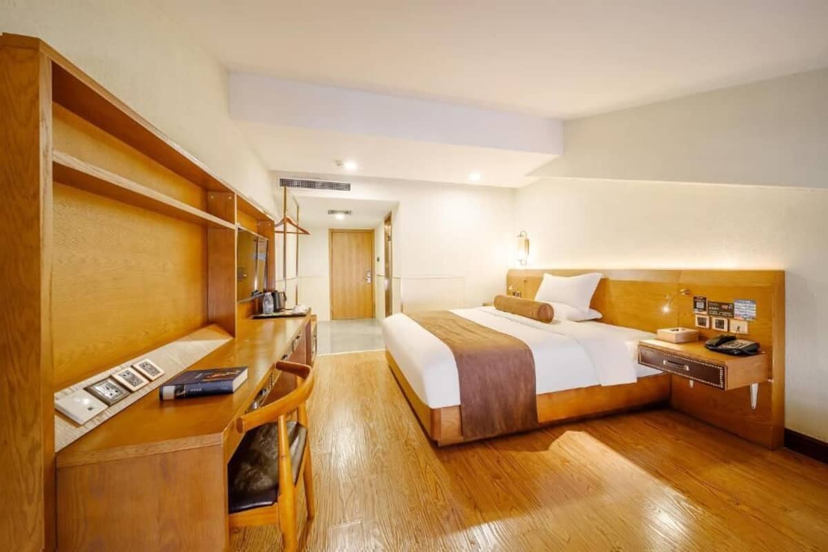 A hotel room with a wooden desk, a bed with a headboard, a bedside table with a lamp, and a wooden wardrobe.