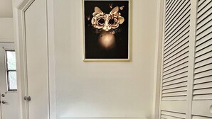 A room with a white door, a framed artwork on the wall, a grey cushioned chair, a small table, and a window.