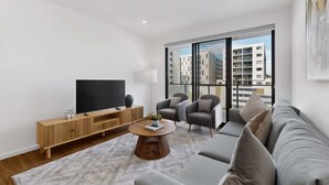 Ein modernes Wohnzimmer mit Flachbildfernseher, Holzkonsole, grauem Sofa und einem großen Fenster mit Blick auf die Stadt.