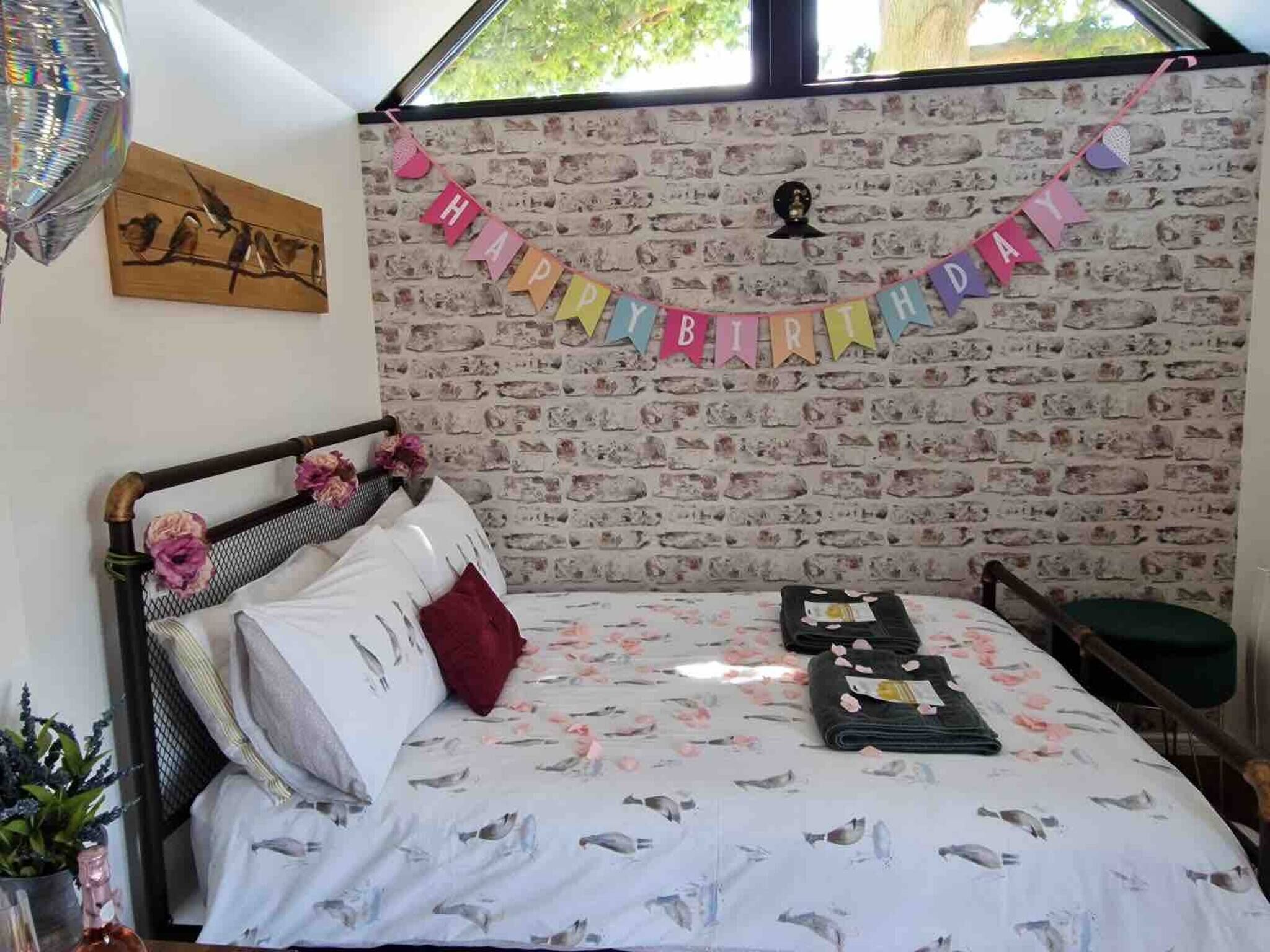 A room with a brick wall, a bed with a floral bedspread, a 'Happy Birthday' banner, and a window with a view of trees.