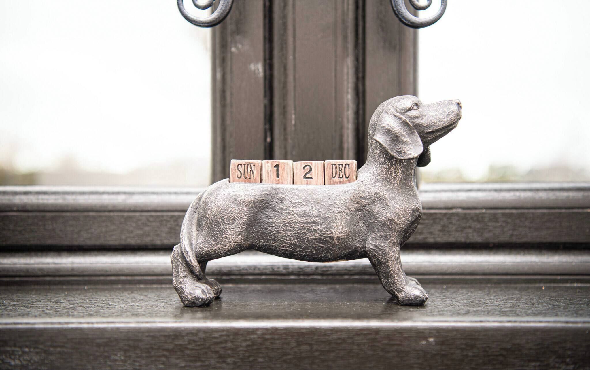 Una figurita de perro de metal con bloques de madera que deletrean 'SUN 12 DEC' sobre un alféizar.