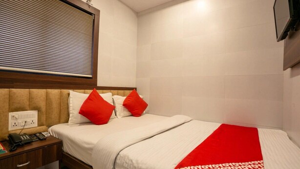 A hotel room with a bed, a nightstand with a phone, a power outlet, and a window with blinds. - Townhouse Oak Ulwe (Panvel)