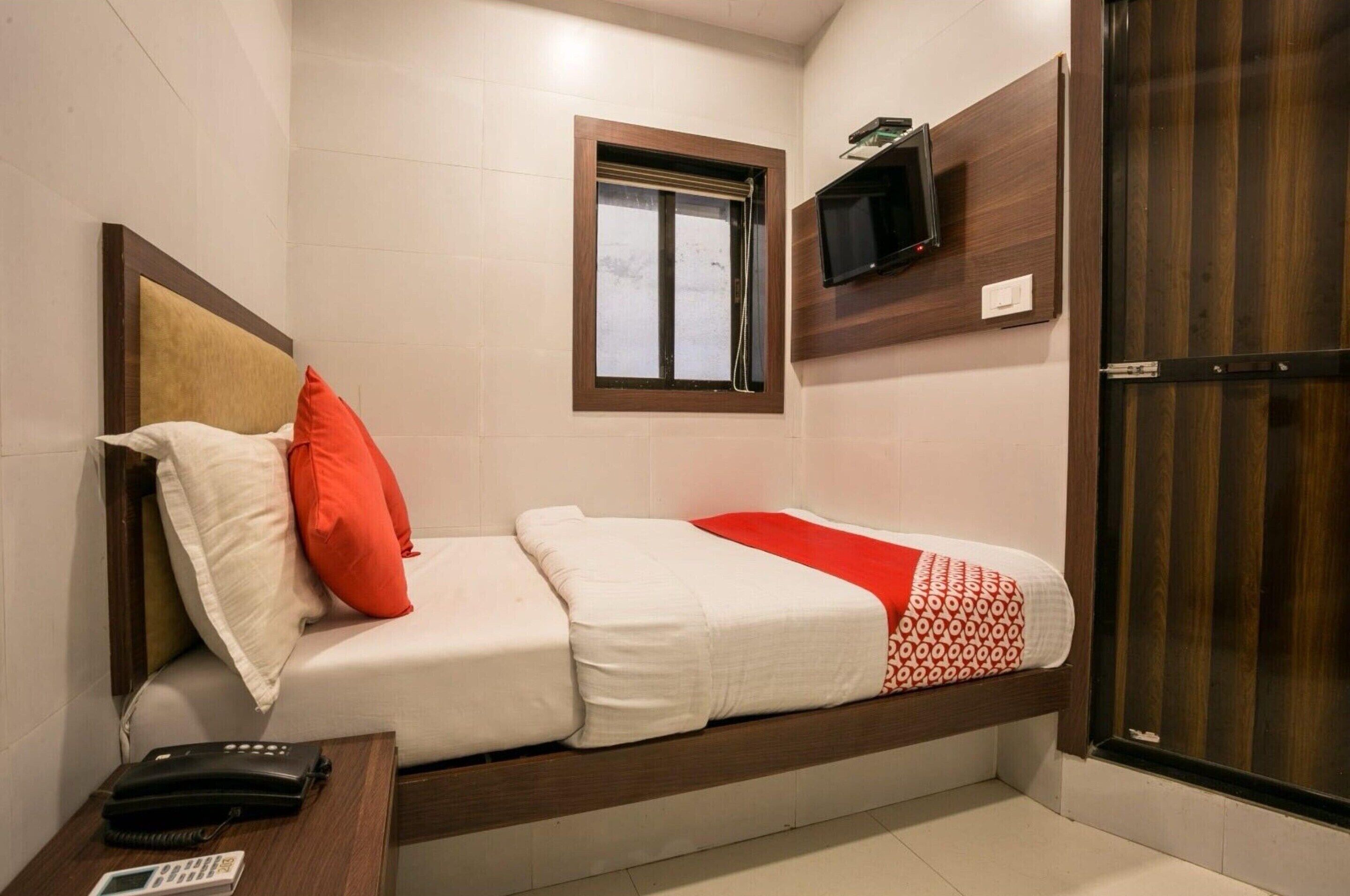 A small, single-bed room with a wooden headboard, a bedside table with a phone, a TV mounted on the wall, and a window with a view.
