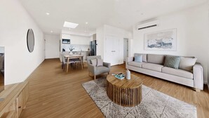 A modern living room with a sofa, coffee table, and a dining area with chairs and a table. - Newly Built Apartment Near Shops and Parks (Cannington)