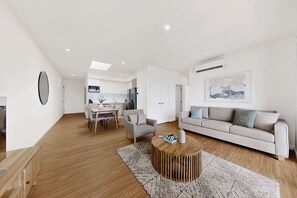 A modern living room with a sofa, coffee table, and a dining area with chairs and a table. - Newly Built Apartment Near Shops and Parks (Cannington)