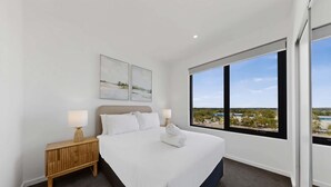 A bedroom with a bed, two bedside lamps, a wooden nightstand, and a large window with a view. - Newly Built Apartment Near Shops and Parks (Cannington)