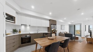 A modern kitchen with white cabinets, a wooden dining table, and a view of the outdoors through a sliding door. - Bright Newly 2 Bed Apartment with Balcony Views (Cannington)