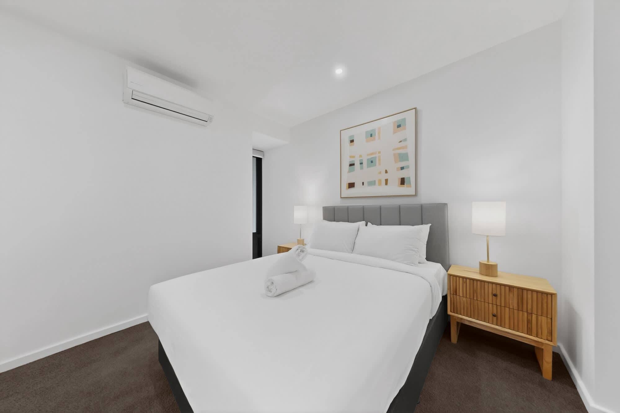 Une chambre d’hôtel moderne avec un grand lit, deux tables de chevet avec des lampes, un climatiseur au plafond et un tableau abstrait encadré accroché au mur.