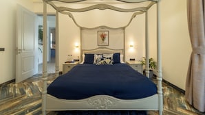 A four-poster bed with a dark blue bedspread, two bedside tables with lamps, a framed picture on the wall, and a door leading to another room. - ELIVAAS Fernweh 4 BHK with Pool (Assagao)