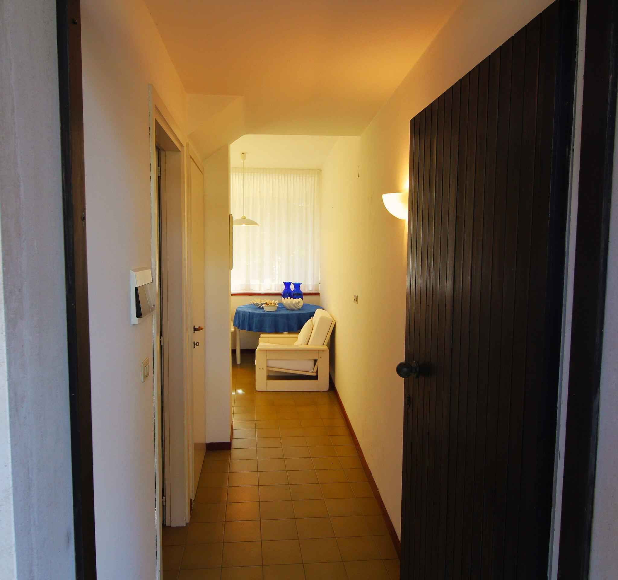 Een smalle gang die leidt naar een slaapkamer met een bed, een stoel en een raam met gordijnen.