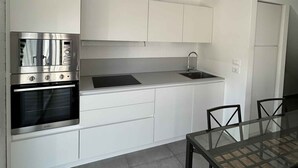 A modern kitchen with white cabinetry, a built-in oven, a sink, and a dining area with a glass table and chairs. - Villa with air conditioning and garden (Lignano Sabbiadoro)