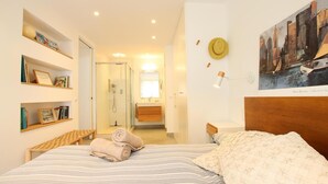 Een hotelkamer met een bed, een badkamer, een stadsgezicht schilderij en een plank met boeken en een hoed.