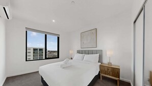 A modern hotel room with a large bed, a nightstand, a wall-mounted lamp, and a view of the city through a large window.