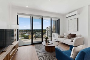 A modern living room with a large sliding glass door, a flat-screen TV, a round coffee table, and a sofa. - Modern,Newly Furnished Apartment with Full Kitchen (Cannington)