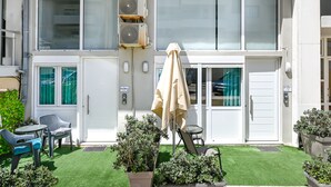 Um edifício com fachada branca, aparelhos de ar condicionado, uma porta com cortina verde e um pequeno pátio com mesa, cadeiras e plantas em vasos.