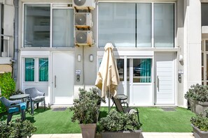 A building with a white exterior, air conditioning units, a door with a green curtain, and a small patio with a table, chairs, and potted plants. - Kalimera Beach Apartment 2 – Town Experience : (Rethymno)