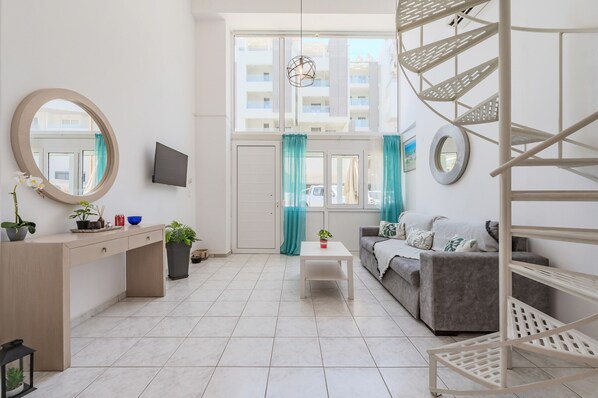 A modern living room with a sofa, a coffee table, a TV, and a staircase. - Kalimera Beach Apartment 1 – Town Experience (Rethymno)
