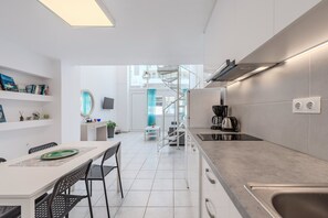 A modern kitchen with white cabinetry, a dining area with a table and chairs, and a staircase leading to an upper level. - Kalimera Beach Apartment 1 – Town Experience (Rethymno)