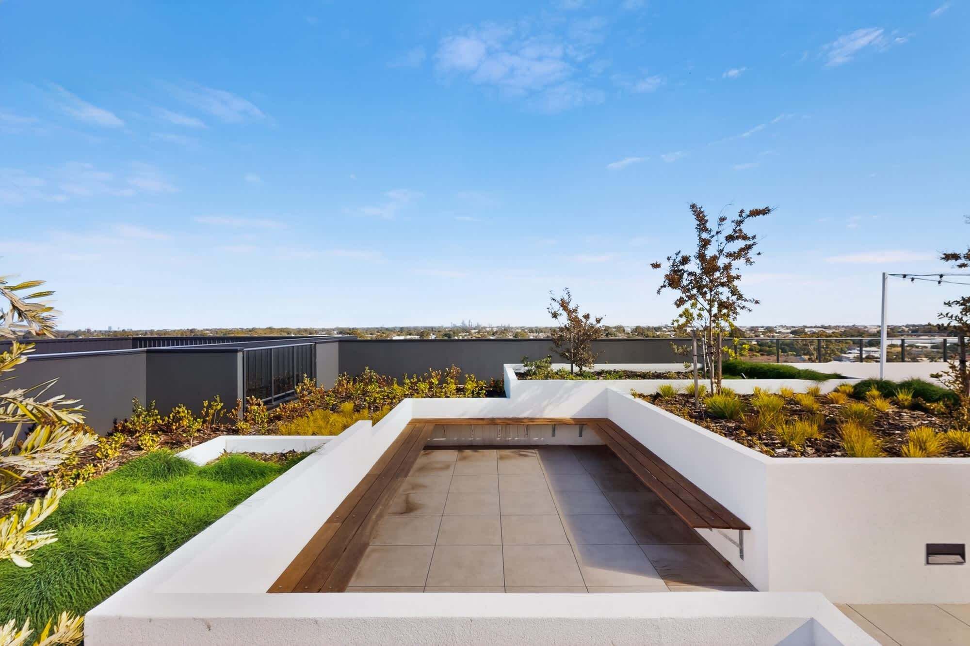 A rooftop terrace with a wooden bench, white walls, and a view of the cityscape.