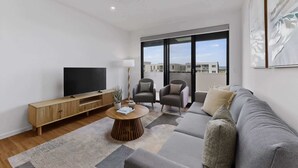 Ein modernes Wohnzimmer mit Flachbildfernseher, Holzmöbeln, einem runden Couchtisch und einem großen Fenster mit Blick auf die Stadt.
