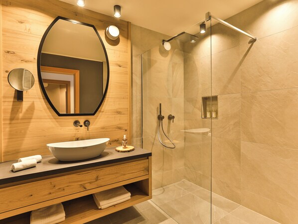Un baño moderno con tocador de madera, un lavabo ovalado grande, un espejo, una ducha con puerta de vidrio y un estante pequeño con artículos de aseo.