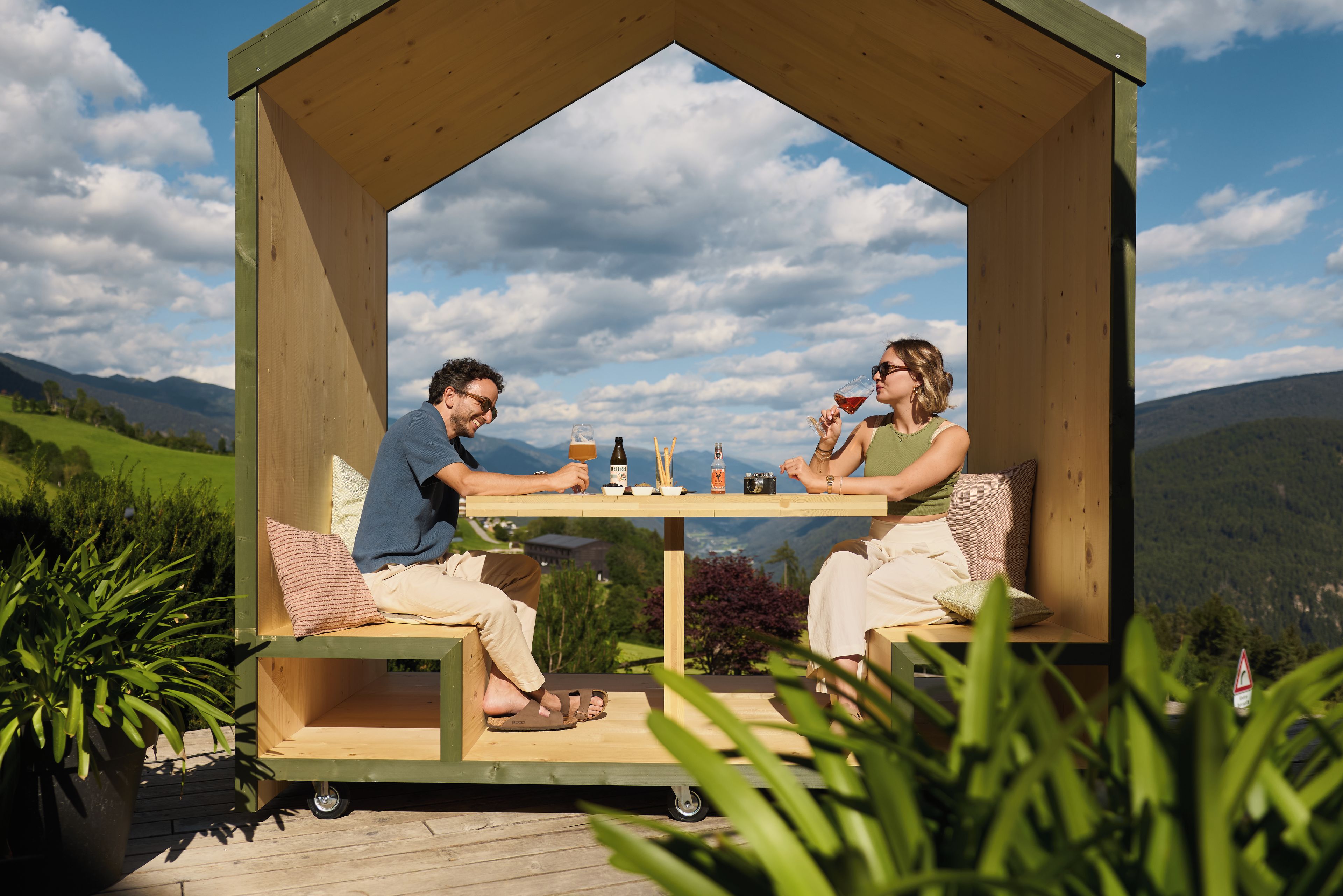 A wooden gazebo with a table and chairs, set against a mountainous backdrop.