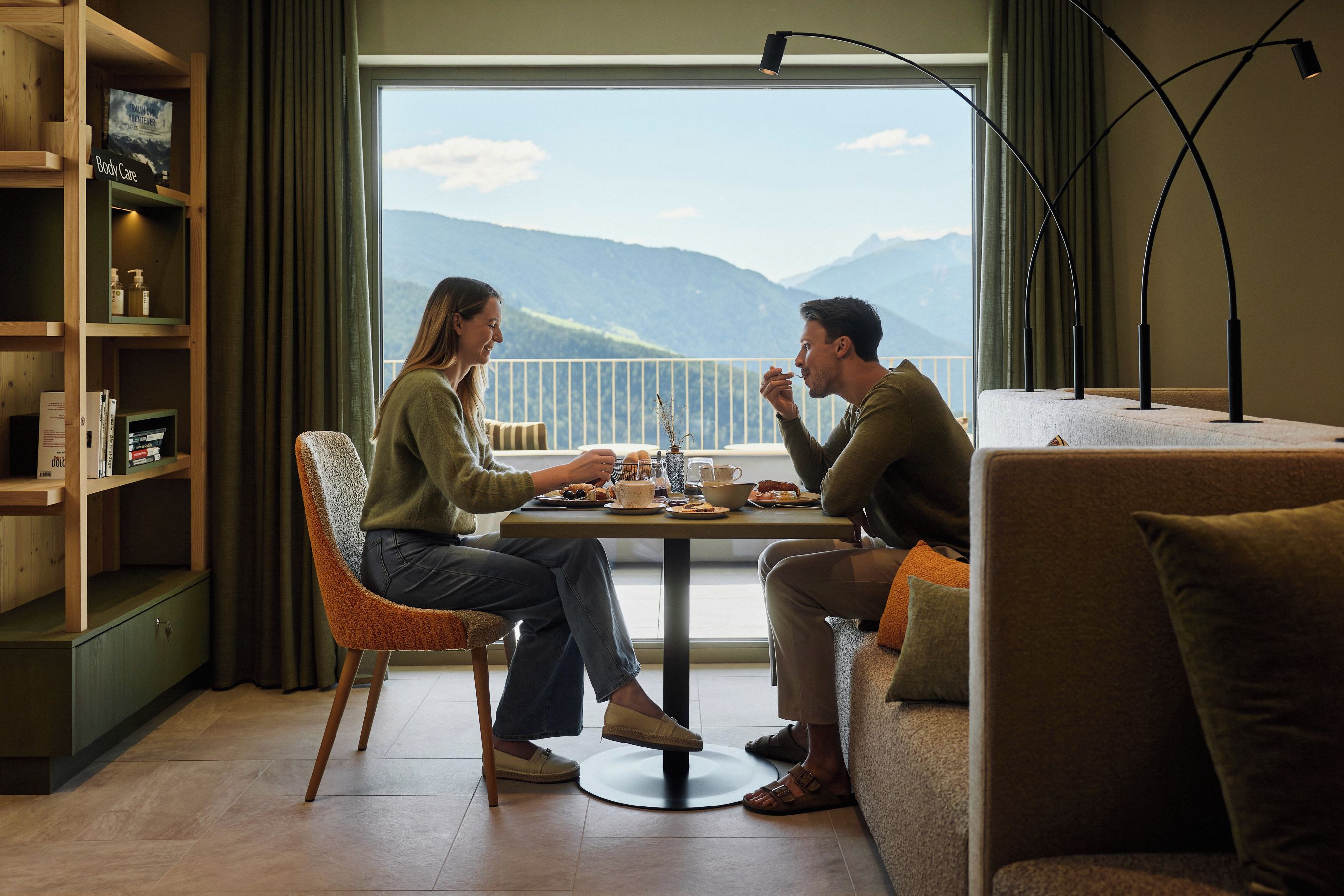 Una habitación con una mesa de comedor lista para dos personas, un sofá, una estantería y vistas a montañas a través de un ventanal grande.