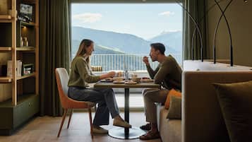 Una habitación con una mesa de comedor lista para dos personas, un sofá, una estantería y vistas a montañas a través de un ventanal grande.