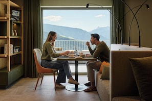 Una habitación con una mesa de comedor lista para dos personas, un sofá, una estantería y vistas a montañas a través de un ventanal grande.