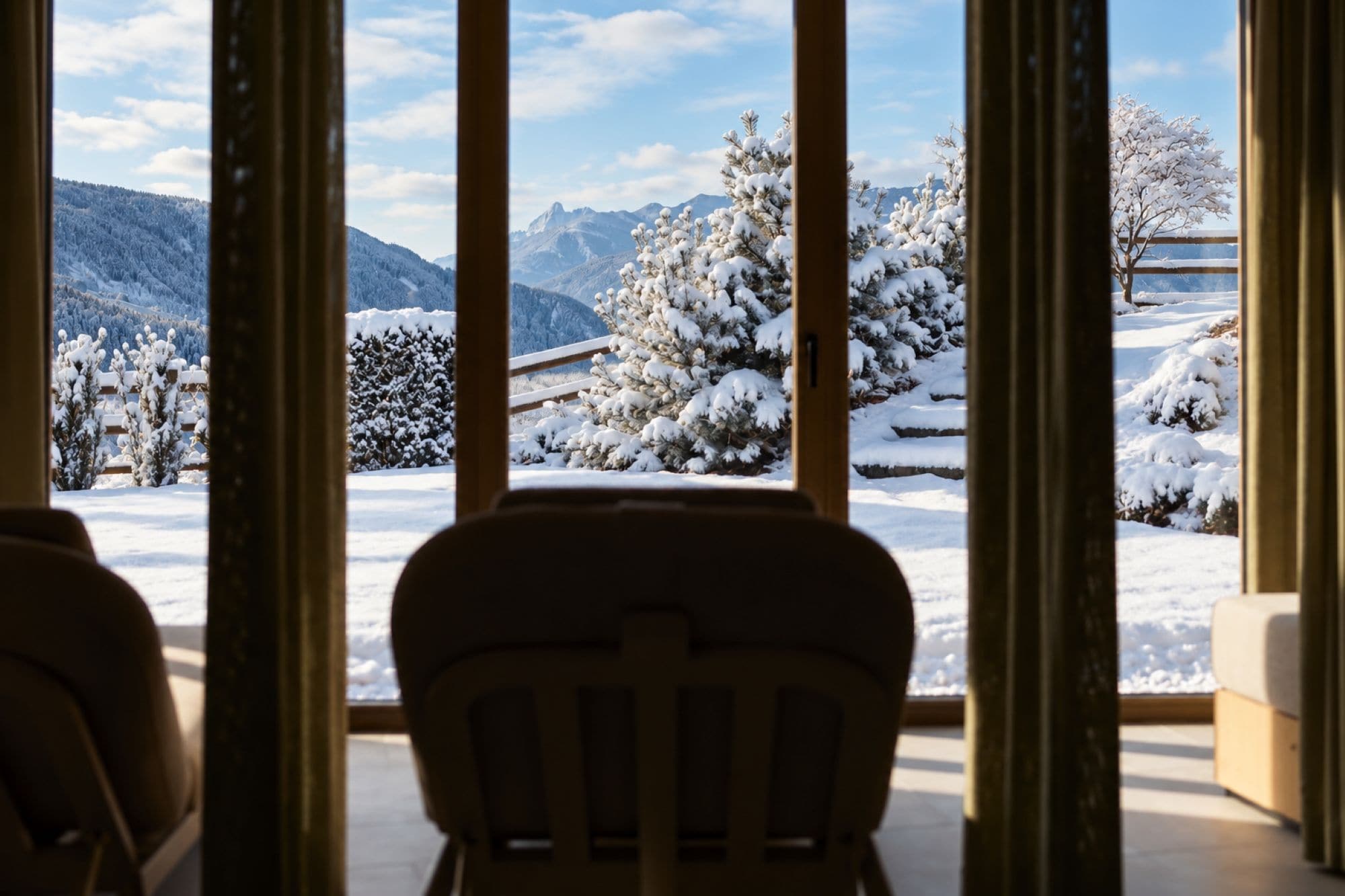 Vista desde una ventana con una silla al frente, mostrando un paisaje nevado con árboles y montañas.